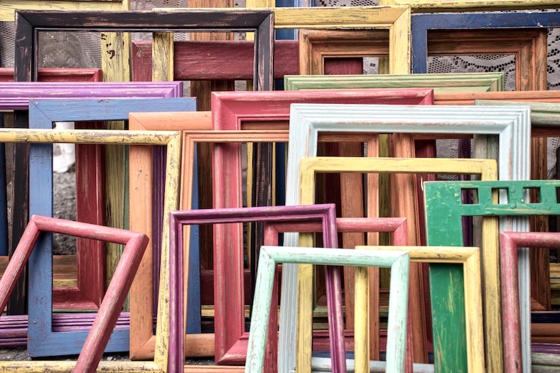 A collected of empty wooden picture frames, painted in a range of colours, leaning up against each other.